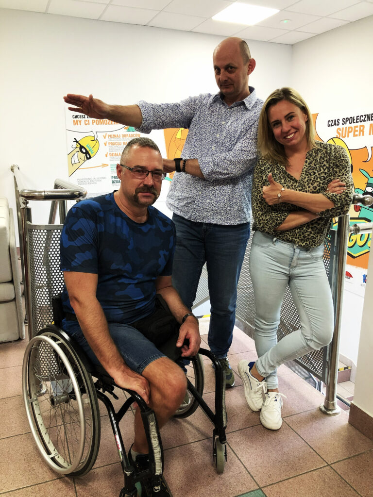 Troje przedstawicieli Bielskiego Stowarzyszenia Artystycznego "Teatr Grodzki" pozuje do zdjęcia w pomieszczeniu 3 sektora we Wrocławiu. Dwóch mężczyzn, jeden z nich jest na wózku dla osoby z niepełnosprawnością, i szeroko uśmiechnięta kobieta. Jeden z mężczyzn unosi ręce w bok.