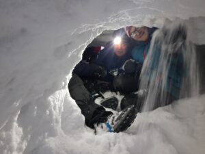 Na zdjęciu dwóch chłopaków siedzących w jamie śnieżnej. z sufitu jamy sypie się śnieg, jeden z chłopaków ma włączoną czołówkę.