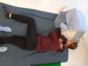 na zdjęciu leżąca osoba w pozycji bocznej ustalonej. Druga osoba - kobieta, nachyla się nad twarzą osoby leżącej i sprawdza czy ta oddycha. Zdjęcie przedstawia ćwiczenia z udzielania pierwszej pomocy.
