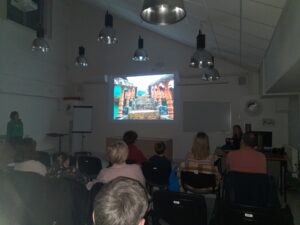 Na zdjęciu sala wykładowa pełna ludzi, którzy słuchają wykładu z pokazem slajdów.