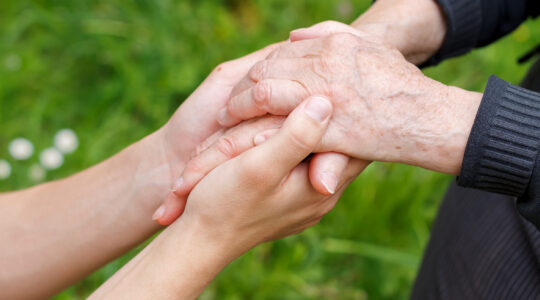 Zdjęcie rąk na tle zielonej trawy. Młodsza osoba obejmuje dłońmi z troską ręce seniorki.