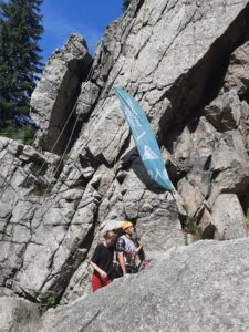 Na zdjęciu dwie osoby idą w górę przy ścianie skalnej. W skałach wbity jest baner BAZY.