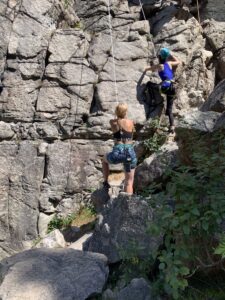 Na zdjęciu młoda dziewczyna zaczyna się wspinać w skałach. Druga kobieta ją asekuruje.