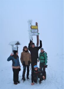 Na zdjęciu grupa 5 osób, które srtoją na szczycie śnieżnika pod znakiem informacyjnym. Jest biało i leży dużo śniegu.