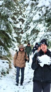 Na zdjęciu grupa osób idzie leśną ściżką w zimie. Chłopak na pierwszym planie trzyma w rękach wielką kulę śniegu.