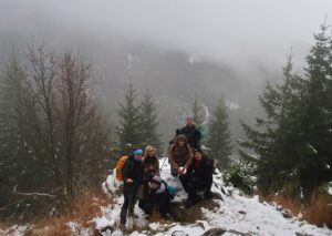 Na zdjęciu grupa 6 osób stoi na skałce pokytej śniegiem. Zanimi w tle góry pokryte lekką mgłą.