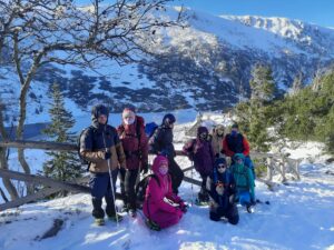 Na zdjęiu grupa osób niewidomych i słabowidzących w zimie w górach. Za nimi w tle góry i zamarznięte jezioro.