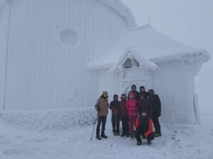 Na zdjęciu grupa ludzi na szczycie Śnieżki w zimie. Za nimi ośnieżony budynek w tle.