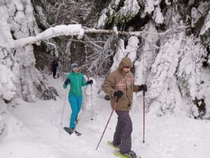 Na zdjęciu dwie osoby w rakietach śnieżnych idą po leśniej ścieżce w zimowych warunkach.