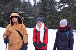 Na zdjęciu 3 młode osoby ubrane w zimowe ciuchy. W tle las w zimowej oprawie.