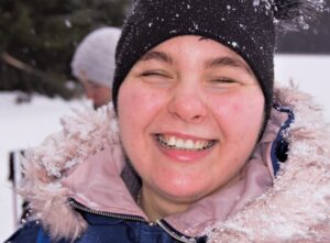Zdjęcie przedstawia twarz młodej uśmiechniętej dziewczyny w czapce i zimowej kurtce. Pada śnieg.