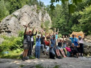 na zdjęciu grupa uśmiechniętyh osób z podniesionymi do góry rękami. W tle ściana skalna i las.