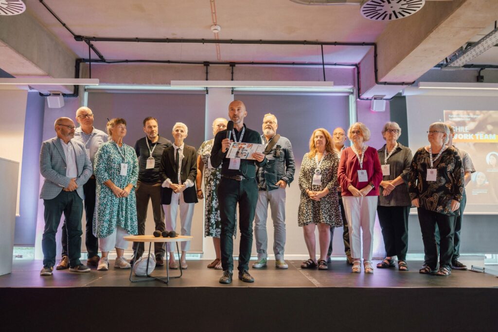 14 osób stoi na scenie w sali konferencyjnej, wśród nich prezes i Fundacji Imago, który przemawia przez mikrofon. Pozostałe osoby to członkowie komitetu stowarzyszenia krótkiej przerwy ISBA. 
