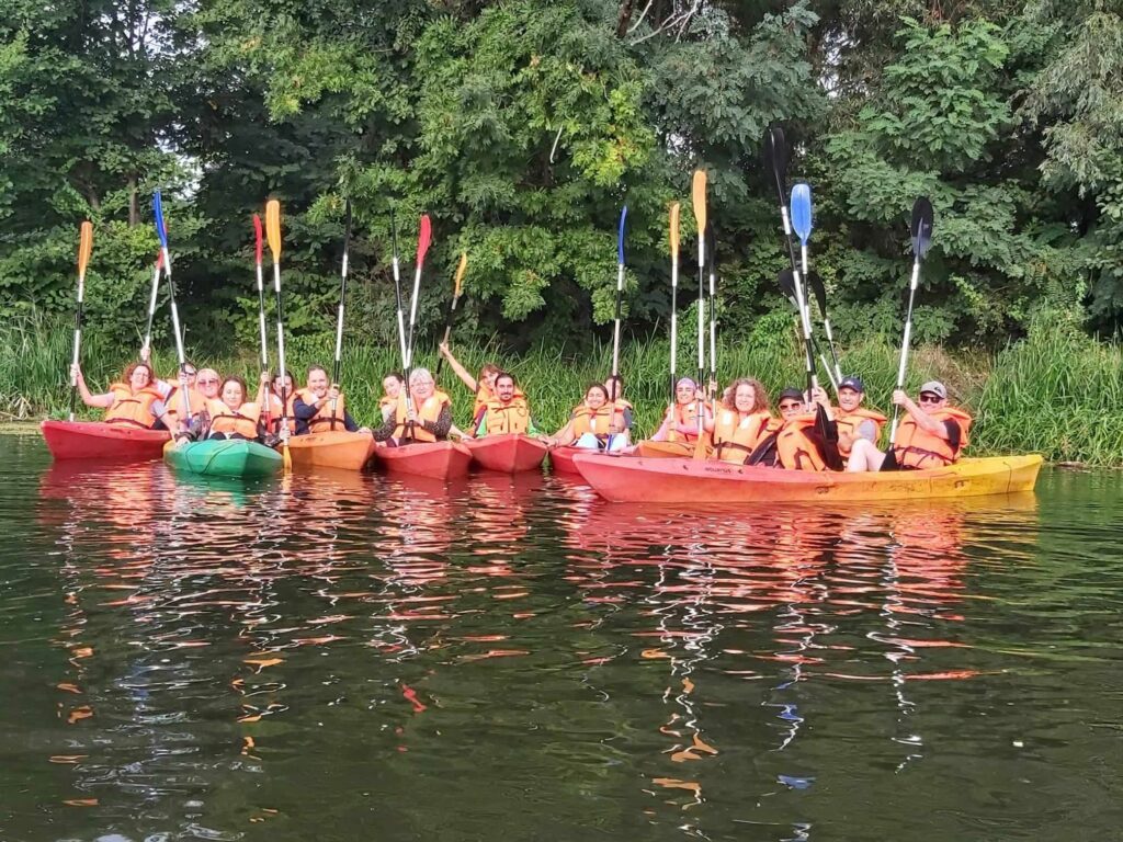 Kilkanaście osób pozuje do zdjęcia na kajakach, są uśmiechnięci, unoszą wiosła w górę. Kajaki są ustawione na wodach rzeki Odry,  jeden obok drugiego. To uczestnicy konferencji ISBA 2023.