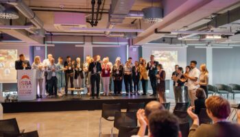 27 osób stoi na scenie, wśród nich prezes i zespół Fundacji Imago oraz komitet stowarzyszenia krótkiej przerwy ISBA. Naprzeciwko nich siedzą uczestnicy konferencji ISBA 2023 we Wrocławiu. Większość osób klaszcze. Wszyscy zgromadzeni są w dużej sali konferencyjnej. To zdjęcie z zakończenia konferencji ISBA 2023.