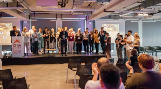 27 osób stoi na scenie, wśród nich prezes i zespół Fundacji Imago oraz komitet stowarzyszenia krótkiej przerwy ISBA. Naprzeciwko nich siedzą uczestnicy konferencji ISBA 2023 we Wrocławiu. Większość osób klaszcze. Wszyscy zgromadzeni są w dużej sali konferencyjnej. To zdjęcie z zakończenia konferencji ISBA 2023.
