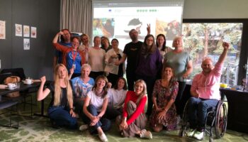 Zdjęcie w sali szkoleniowej projektu "Mam Tę Moc". Na zdjęciu jest 17 osób, uśmiechają się i unoszą ręce w górę, pozując do zdjęcia. Są to uczestnicy, prowadzący, eksperci biorący udział w projekcie "Mam Tę Moc".