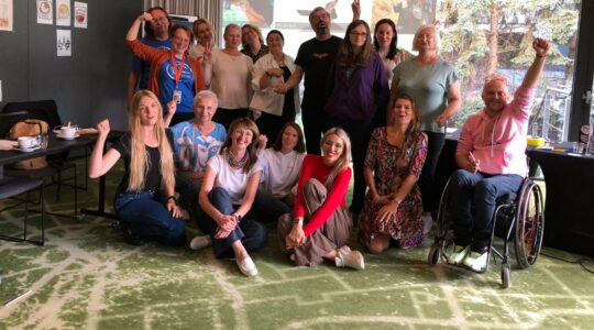 Zdjęcie w sali szkoleniowej projektu "Mam Tę Moc". Na zdjęciu jest 17 osób, uśmiechają się i unoszą ręce w górę, pozując do zdjęcia. Są to uczestnicy, prowadzący, eksperci biorący udział w projekcie "Mam Tę Moc".