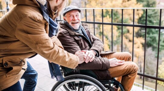 Zdjęcie straszego, roześmianego mężczyzny na wózku. Obok młodszy mężczyzna, asystent, podaje mu rękę.