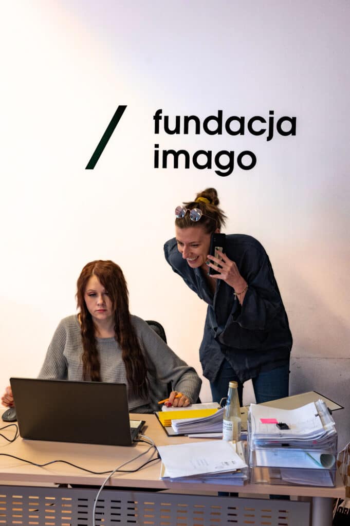 Dwie kobiety w biurze Imago (na ścianie w tle widać logotyp Fundacji Imago). Jedna z nich siedzi przy biurku, przed komputerem, obok leżą dokumenty. Druga pochyla się nad nią, rozmawiając przez telefon.