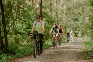Na zdjęciu osoby jadące na rowerach jeden za drugim leśną ścieżką