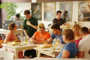 Na zdjęciu grupa ludzi siedząca przy stłach w restauracji. Ludzie rozmawiają ze sobą, dyskutują i jedzą.