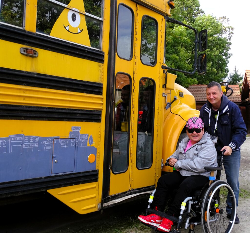 Dwie osoby pozują do zdjęcia obok żółtego pojazdu - autobusu. Jedna z nich jest na wózku dla osoby z niepełnosprawnością. Uśmiechają się.