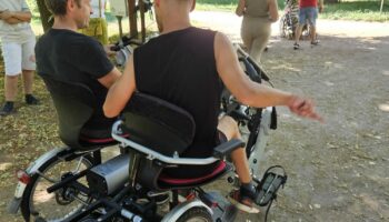 Zdjęcie wykonane w parku. Rower tandem, w którym siedzenia umieszczone są obok siebie. Na siedzeniach siedzą dwie osoby, jedna z nich ma niepełnosprawność ruchową.