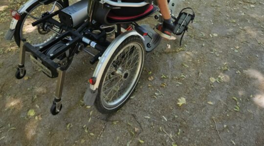 Zdjęcie wykonane w parku. Rower tandem, w którym siedzenia umieszczone są obok siebie. Na siedzeniach siedzą dwie osoby, jedna z nich ma niepełnosprawność ruchową.