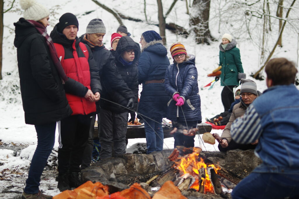 Grupa uczestników spotkania w Lasu Osobowickim stoi/siedzi przy ognisku, pieką kiełbaski i serki. 