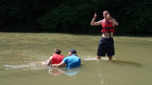 Na zdjęciu 3 mężczyzn. 2 z nich jest zanurzonycch w wodze, jeden stoi w wodzie po kolana i pokazuje oba kciuki do góry