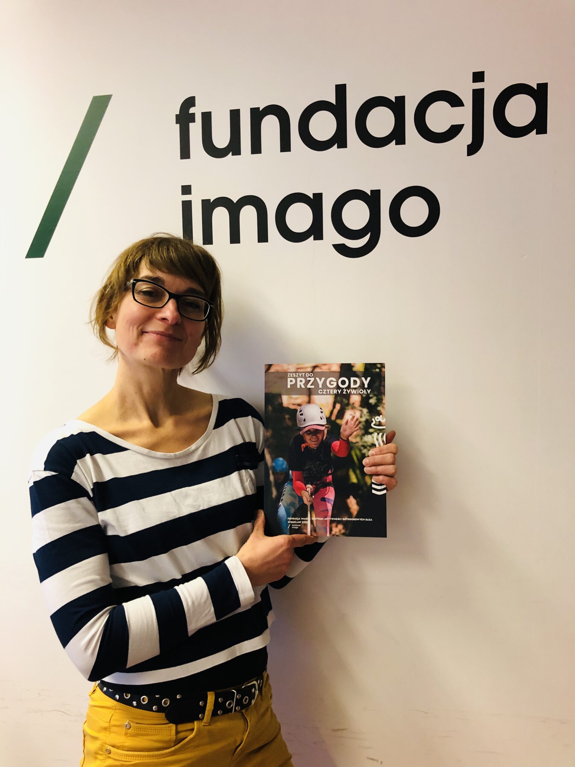 Uśmiechnięta Magdalena Stempska z Fundacji Imago prezentuje okładkę publikacji „Zeszyt do przygody”. Fotografia na tle białej ściany z logotypem Fundacji Imago.