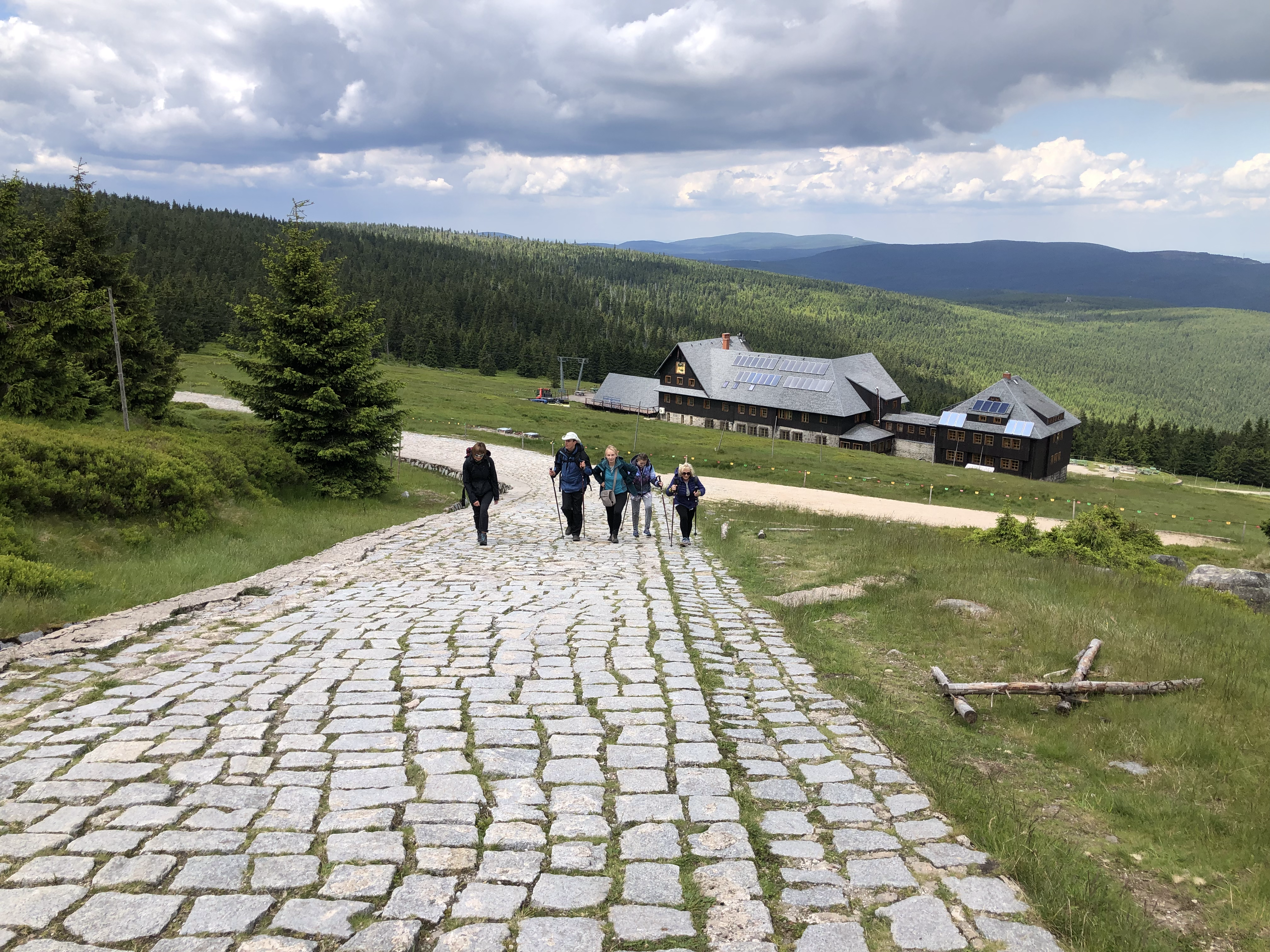 Wycieczka górska. Grupa kilku osób idzie pod górę, wybrukowaną ścieżką. W tle góry i schronisko.