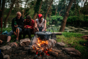 Na zdjęciu gotujący się kociołek na ogniu, w tle grupa 3 osób siedzi na drewnianej ławce
