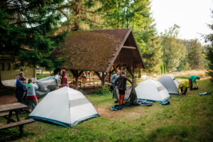Na zdjęciu osoby rokładające namioty na polu kempingowym obok wiaty leśnej