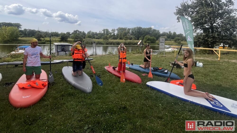 Uczestnicy spotkania nad wodą, stoją przy brzegu, przy deskach SUP, niektórzy mają kamizelki ratunkowe. Naprzeciwko nich stoi instruktorka SUP, która wyjaśnia zasady pływania na deskach.