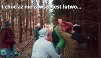 Kilka osób w lesie, w czasie warsztatu dotyczącego terapii dzikością. Są w trakcie wykonywania ćwiczenia z pajęczą siecią (wykonaną z liny). Na górze napis: "I chociaż nie zawsze jest łatwo...".