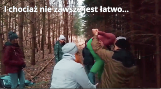 Kilka osób w lesie, w czasie warsztatu dotyczącego terapii dzikością. Są w trakcie wykonywania ćwiczenia z pajęczą siecią (wykonaną z liny). Na górze napis: "I chociaż nie zawsze jest łatwo...".