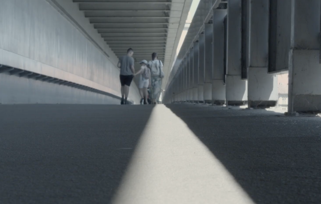 Zdjęcie trzech osób - dorosłego mężczyzny oraz kobiety i dziewczynki idących razem przez parking lub podobną przestrzeń. Mężczyzna trzyma dziewczynkę za rękę. Są odwróceni plecami do osoby robiącej zdjęcie.