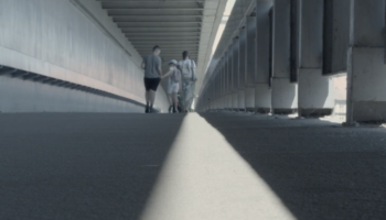 Zdjęcie trzech osób - dorosłego mężczyzny oraz kobiety i dziewczynki idących razem przez parking lub podobną przestrzeń. Mężczyzna trzyma dziewczynkę za rękę. Są odwróceni plecami do osoby robiącej zdjęcie.