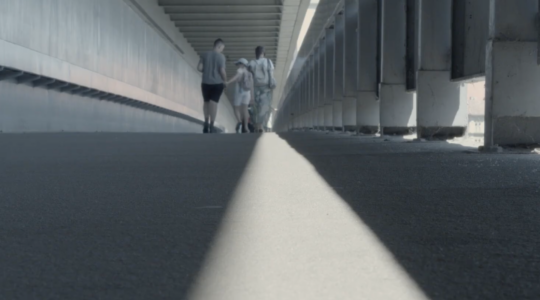 Zdjęcie trzech osób - dorosłego mężczyzny oraz kobiety i dziewczynki idących razem przez parking lub podobną przestrzeń. Mężczyzna trzyma dziewczynkę za rękę. Są odwróceni plecami do osoby robiącej zdjęcie.