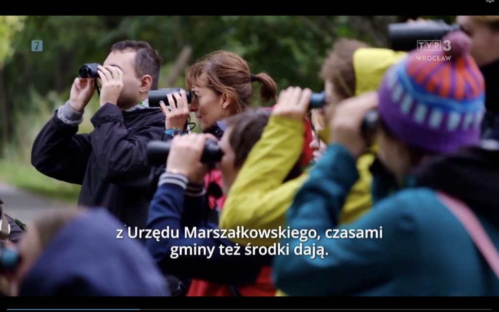 Skrin z ekranu komputera - kadr z programu "Magazyn Dolnośląski". Na zdjęciu grupa osób w czasie wycieczki. Osoby na zdjęciu mają na sobie sportowe ubrania, przy oczach trzymają lornetki i zajęte są obserwacją przyrody.