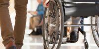Wózek dla osoby z niepełnosprawnością. Widać koła i rękę człowieka, który korzysta z wózka. Obok idzie inna osoba, na zdjęciu widoczne są jej nogi.