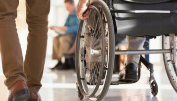 Wózek dla osoby z niepełnosprawnością. Widać koła i rękę człowieka, który korzysta z wózka. Obok idzie inna osoba, na zdjęciu widoczne są jej nogi.