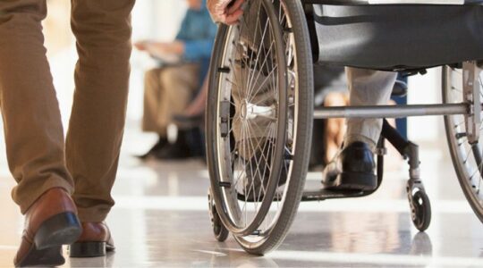 Wózek dla osoby z niepełnosprawnością. Widać koła i rękę człowieka, który korzysta z wózka. Obok idzie inna osoba, na zdjęciu widoczne są jej nogi.