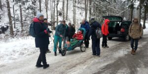 Na zdjęciu grupa osób przygotowuje się do górskiej wędrówki - jedna z nich jest na wózku trekkingowym. w tle las i otwarty bagażnik samochodu.