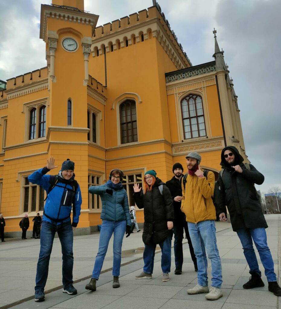 Druga grupa szkoleniowa przed budynkiem dworca we Wrocławiu. Przed budynkiem dworca stoi sześć osób, machają do zdjęcia, uśmiechają się. Magda Stempska z Fundacji Imago ma na uszach słuchawki wyciszające - uczestnicy wcielali się w osoby Głuche.