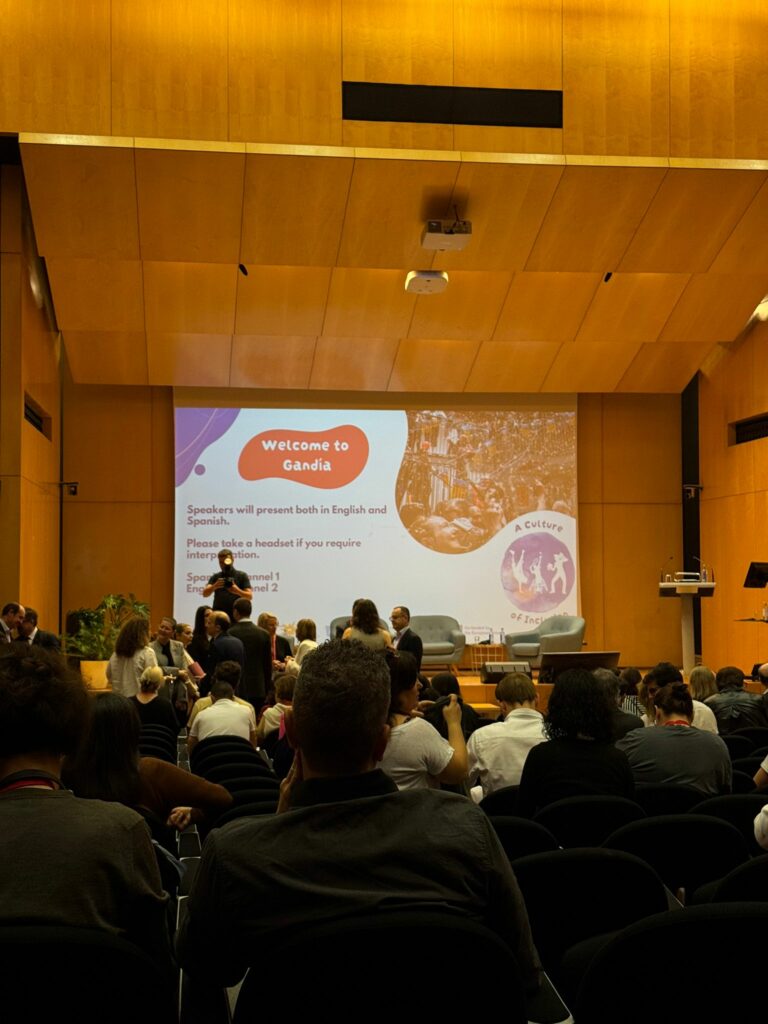 Sala konferencyjna, pod ścianą duży ekran z prezentacją, przed nim prelegent. Naprzeciwko słuchacze siedzący na krzesłach, w sali jest dużo osób.