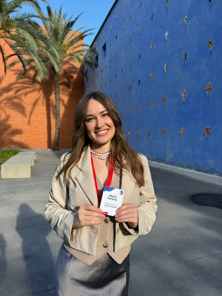 Uśmiechnięta Mariia Pavlova z Fundacji Imago pozuje do zdjęcia przed budynkami, gdzie odbywa się konferencja, w rękach trzyma konferencyjny identyfikator.
