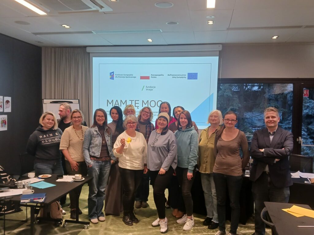 Uczestniczki, uczestnicy i prowadząca grupę roboczą Mam tę moc w sali szkoleniowej na tle ekranu z napisem "Mam tę moc" i logotypami szkoleniowymi.